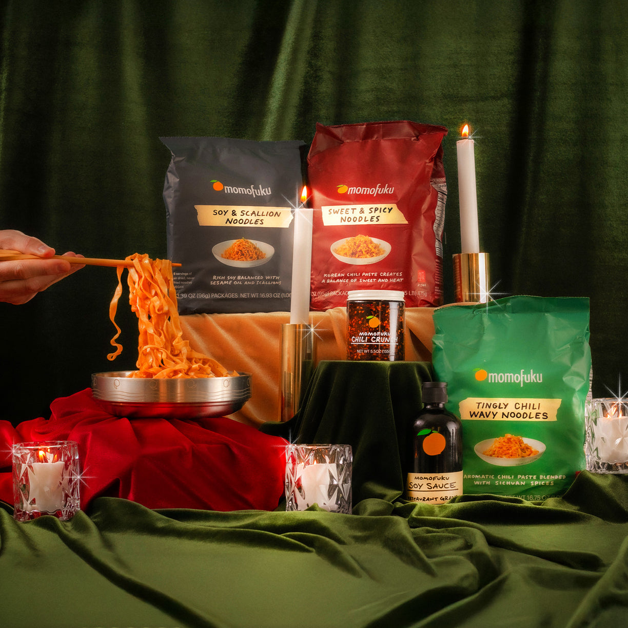 Festive display of 3 Momofuku Noodle flavors, 1 jar of Chili Crunch, 1 bottle of Soy Sauce, and chopsticks pulling noodles out of a Momofuku Noodle Bowl on velvet green background