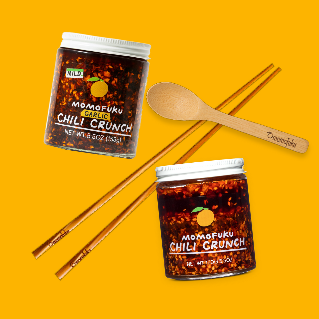 Two jars of Momofuku Chili Crunch on a yellow background with wooden spoon and chopsticks.