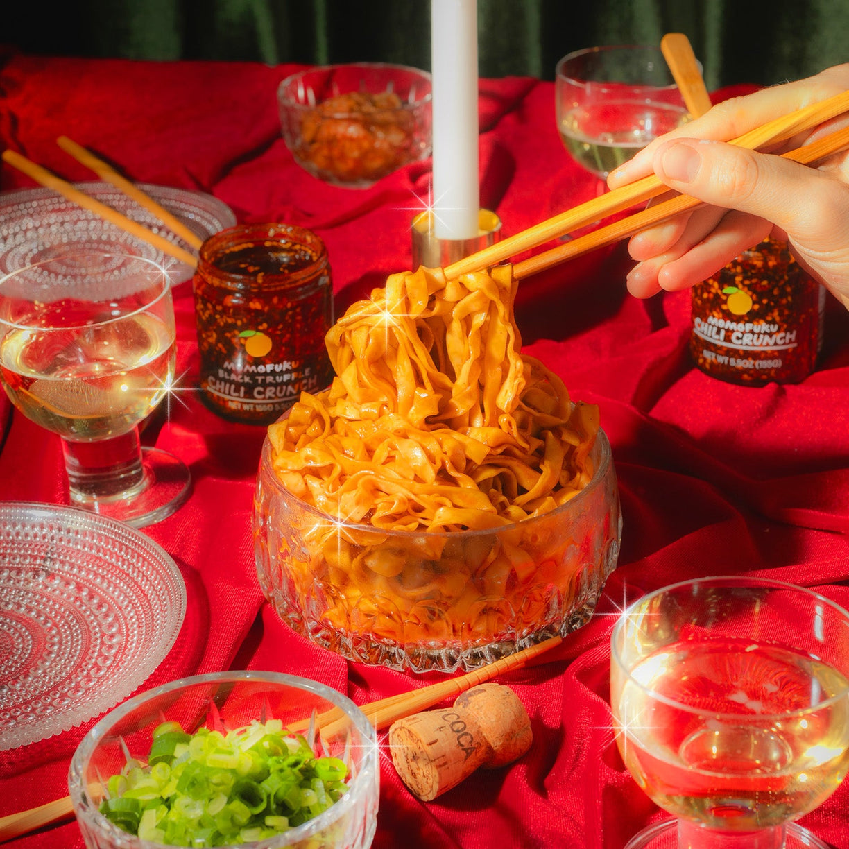 festive tablescape with 2 jar of chili crunches, and noodles being pulled by chopsticks in glass bowl